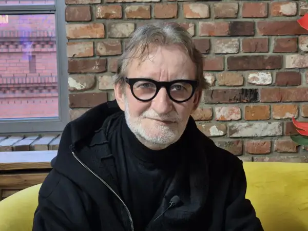 Maciej Mańkowski w okularach, z siwą brodą, w czarnej bluzie na tle ceglanej ściany.