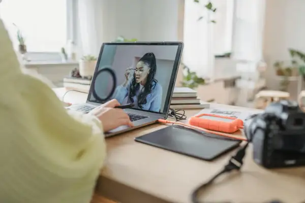 Osoba pracuje przy laptopie, poprawiając makijaż na zdjęciu. W tle widać aparat i dysk zewnętrzny, sugerując pracę nad rozjaśnianiem zdjęć.